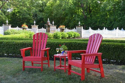 Two red Adirondack chairs with a small table, set in a garden with greenery and a white picket fence in the background.