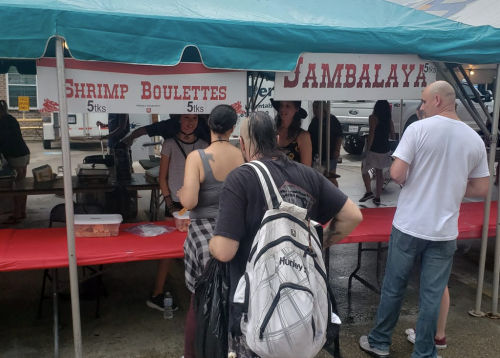 A food stall at a market offers shrimp boulette and jambalaya, with customers interacting under a tent.