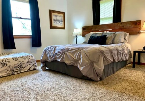 Cozy bedroom with a queen bed, decorative pillows, a wooden headboard, and soft carpet. Natural light from a window.