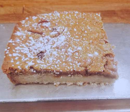 A slice of dessert with a golden-brown top, dusted with powdered sugar, on a silver tray.