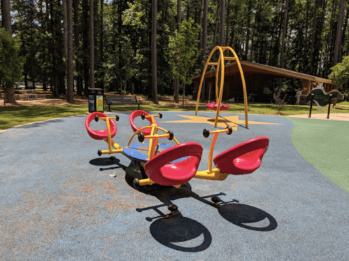 Colorful playground equipment with a seesaw and swings, surrounded by trees in a sunny park.