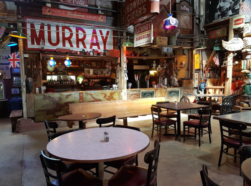 A rustic bar interior with colorful signs, wooden tables, and a long counter, creating a lively, vintage atmosphere.