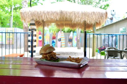 A burger with toppings and fries on a tray, set on a colorful outdoor table with a thatched roof in the background.