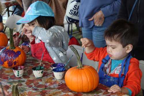 Children painting pumpkins at a table, with colorful paints and decorated pumpkins in a festive setting.