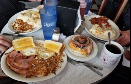 A table with plates of breakfast food, including eggs, hash browns, toast, ham, and a cinnamon roll, with drinks.
