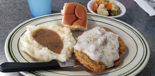 A plate with fried chicken smothered in gravy, mashed potatoes, a roll, and a side of mixed vegetables.