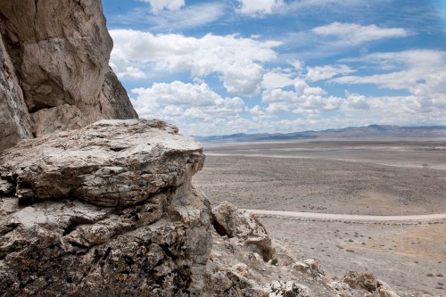 A rocky outcrop overlooks a vast, arid landscape under a cloudy sky, with distant mountains on the horizon.