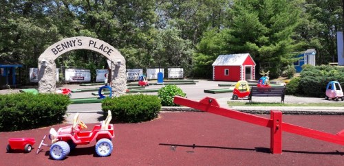 A colorful playground with a stone archway labeled "Benny's Place," featuring toy cars, a seesaw, and a red playhouse.