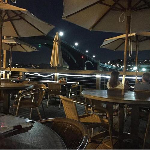 A nighttime view from a restaurant patio, featuring umbrellas and a bridge illuminated in the background.