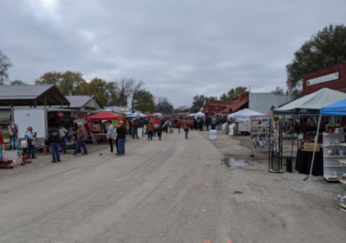 A bustling outdoor market scene with vendors, tents, and visitors walking along a gravel path under a cloudy sky.