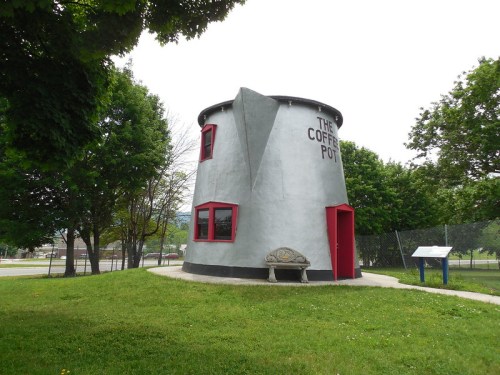 A quirky, cylindrical building resembling a coffee pot, with red accents and surrounded by green grass and trees.