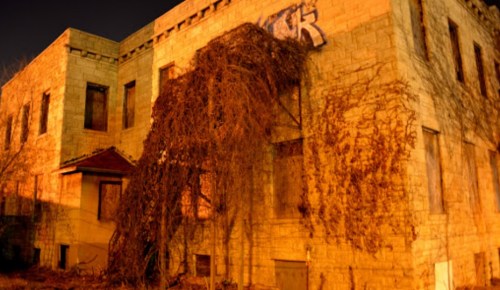 Abandoned building at night, covered in vines, with graffiti on the stone walls and boarded-up windows.