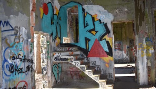 Graffiti-covered walls and a staircase in a dilapidated, abandoned building. Sunlight filters through the openings.