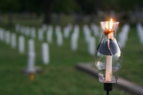 A lit candle in a glass holder stands in a cemetery, with white markers visible in the background.