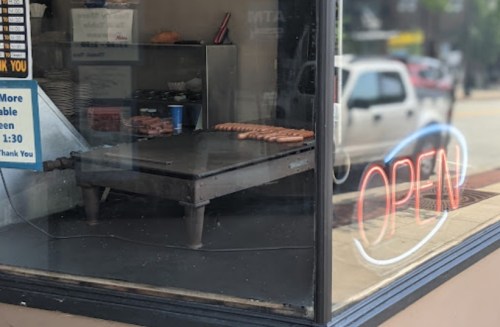 A storefront with an "OPEN" neon sign, featuring a grill with hot dogs cooking inside.