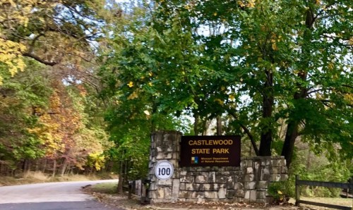 Sign for Castlewood State Park surrounded by trees with autumn foliage, near a winding road.