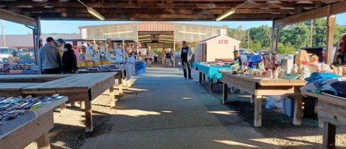 A bustling market with vendors and shoppers, featuring tables filled with various goods under a wooden structure.