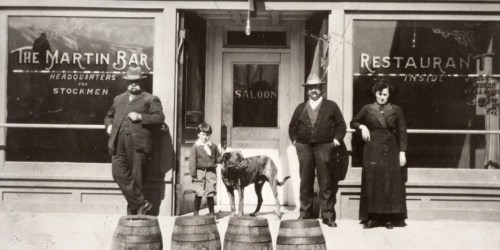 A historic black-and-white photo of four people and a dog outside a saloon, with barrels in front.