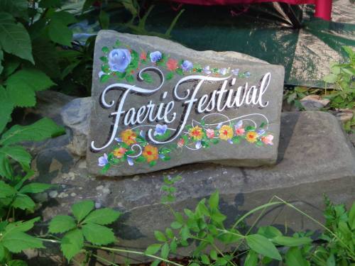 A painted stone sign reading "Faerie Festival," surrounded by green foliage and flowers.