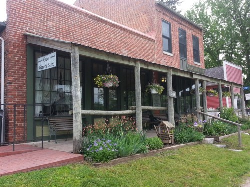A historic general store with a porch, surrounded by greenery and flowers, showcasing a charming rural setting.