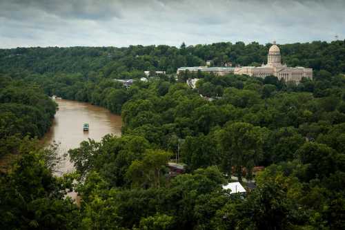 A river flows through lush greenery, with a large building visible on a hill in the background under a cloudy sky.