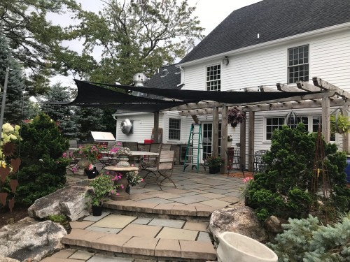 A backyard patio with a stone floor, wooden pergola, black shade cloth, and various plants and seating arrangements.