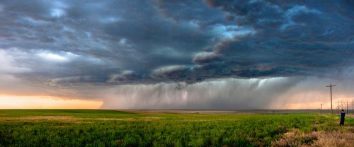 A dramatic sky filled with dark clouds and rain, overlooking a green field and power lines in the distance.