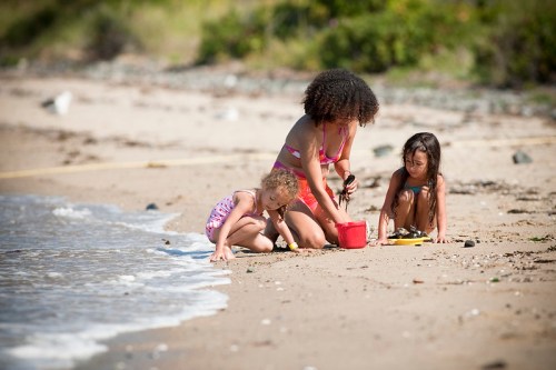A woman and two young girls play in the sand by the beach, enjoying a sunny day near the water.