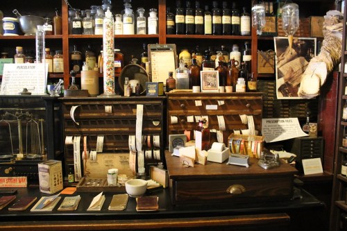 A vintage apothecary shop with shelves of jars, bottles, and a wooden counter filled with old prescription records.