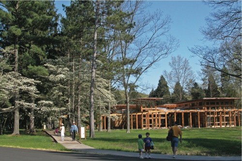 A modern wooden building surrounded by trees, with people walking on a path in a sunny park setting.