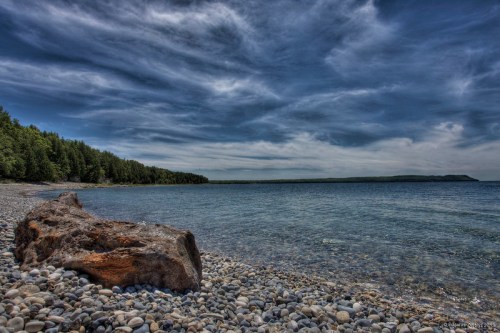 A rocky shoreline with a large log, clear water, and a cloudy sky over a distant green landscape.