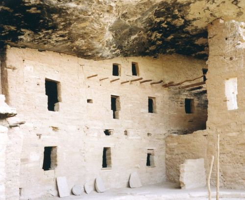 Ancient stone ruins with windows and wooden beams, set under a rocky overhang.
