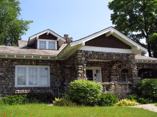 A charming stone house with a gabled roof, surrounded by greenery and colorful flowers under a clear blue sky.