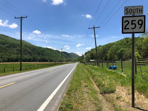 A rural road with a "South 259" sign, flanked by green hills and power lines under a blue sky with clouds.