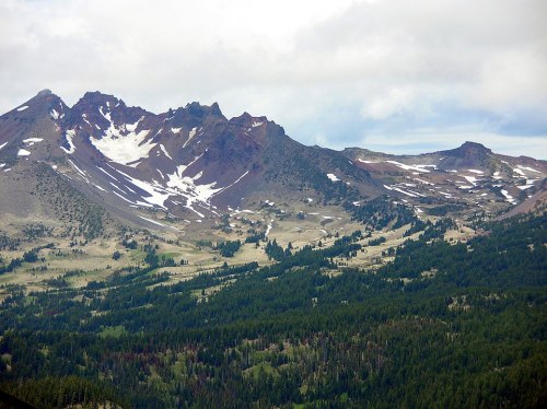 A scenic view of rugged mountains with patches of snow, surrounded by lush green forests under a cloudy sky.