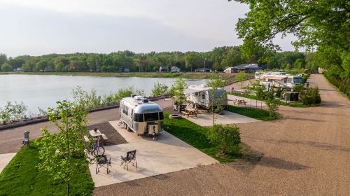 A scenic view of a lakeside RV park with several trailers, picnic areas, and green trees under a clear sky.