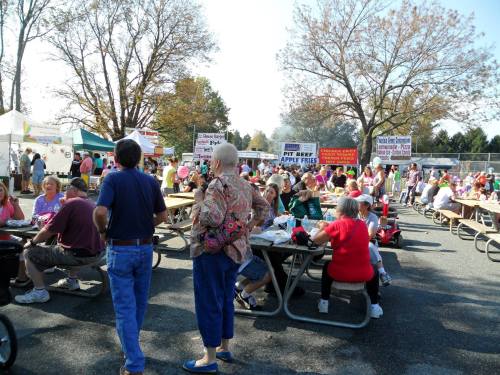 A lively outdoor festival with people enjoying food at picnic tables, surrounded by colorful vendor signs and trees.