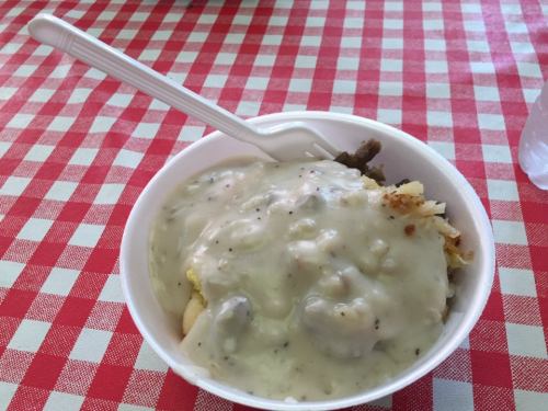 A bowl of creamy gravy over a bed of potatoes and meat, served with a plastic fork on a red and white checkered tablecloth.