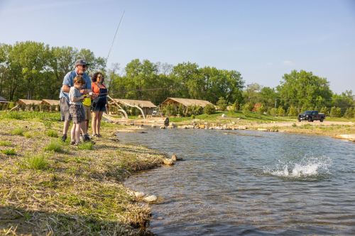A family fishing by a pond, with one person casting a line and water splashing nearby. Tents and trees in the background.