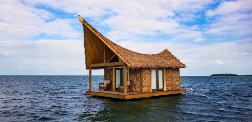 A thatched-roof hut on stilts floating in calm water, surrounded by a blue sky with scattered clouds.