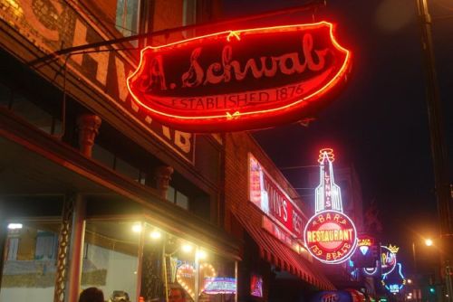 Neon signs for A. Schwab and other establishments illuminate a lively street at night.