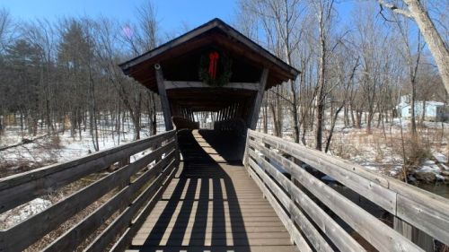 A covered wooden bridge with a festive wreath, surrounded by snowy trees and a clear blue sky.