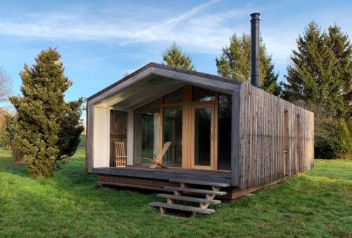 A modern wooden cabin with large windows, a slanted roof, and a chimney, surrounded by greenery.
