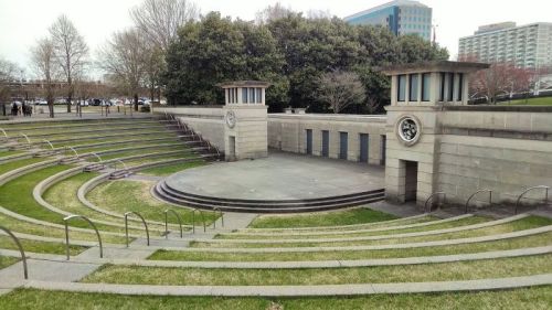 An outdoor amphitheater with tiered seating, surrounded by greenery and buildings in the background.