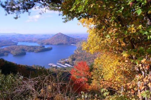 A scenic view of a lake surrounded by autumn foliage and rolling hills under a clear blue sky.