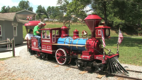 A colorful miniature steam train with flags, parked on a gravel track in a green park setting.