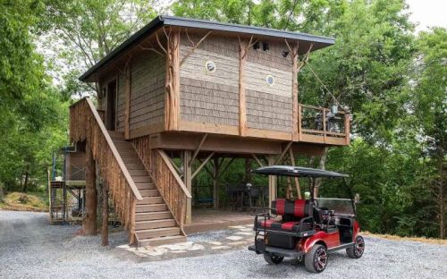 A treehouse with wooden siding and a staircase, beside a golf cart parked on a gravel driveway, surrounded by trees.