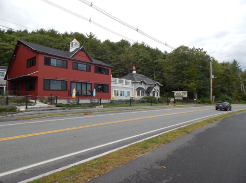 A red and white building along a road, surrounded by trees, with a car driving by on a cloudy day.
