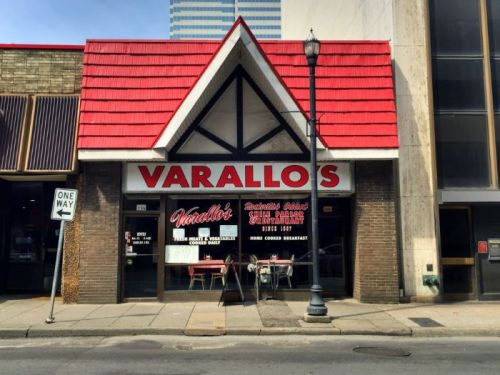 Front view of Varallo's restaurant, featuring a peaked roof and outdoor seating, with a city skyline in the background.