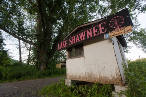 A small, weathered kiosk with "Lake Shawnee" sign, surrounded by greenery along a gravel road.